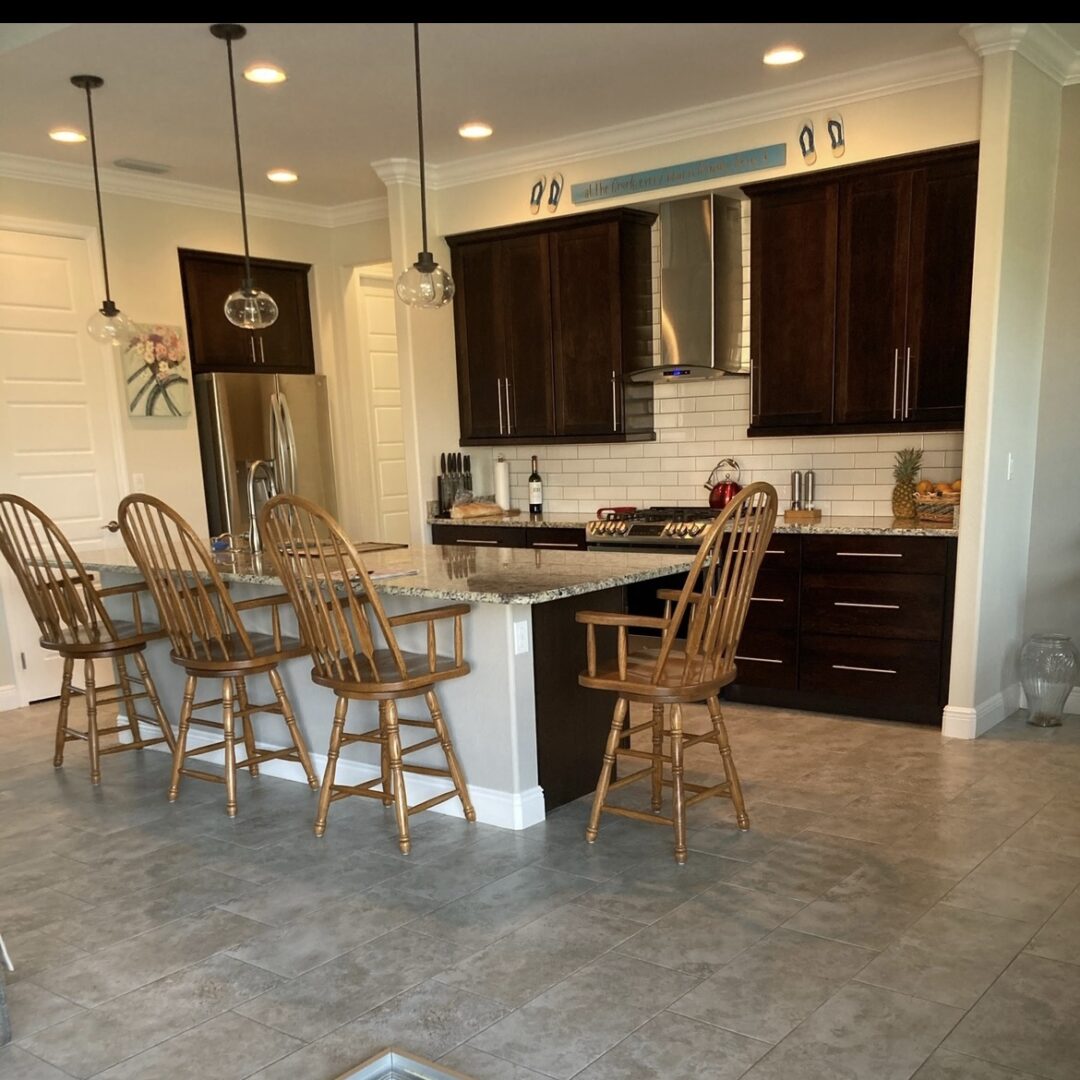 A modern kitchen with dark wooden cabinets, stainless steel appliances, and a large island with a granite countertop. The space is accented by wooden barstools, pendant lighting, and tiled flooring for a clean, inviting atmosphere.