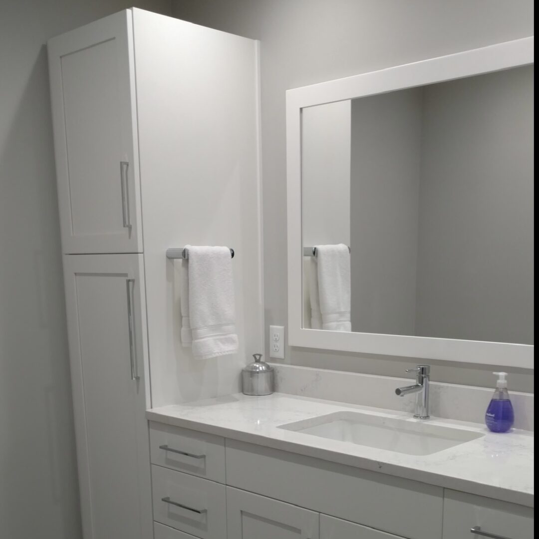 A bright and tidy bathroom area with a white vanity, a framed mirror, and soft lighting. The countertop includes a silver dispenser and a purple soap bottle for a subtle pop of color.