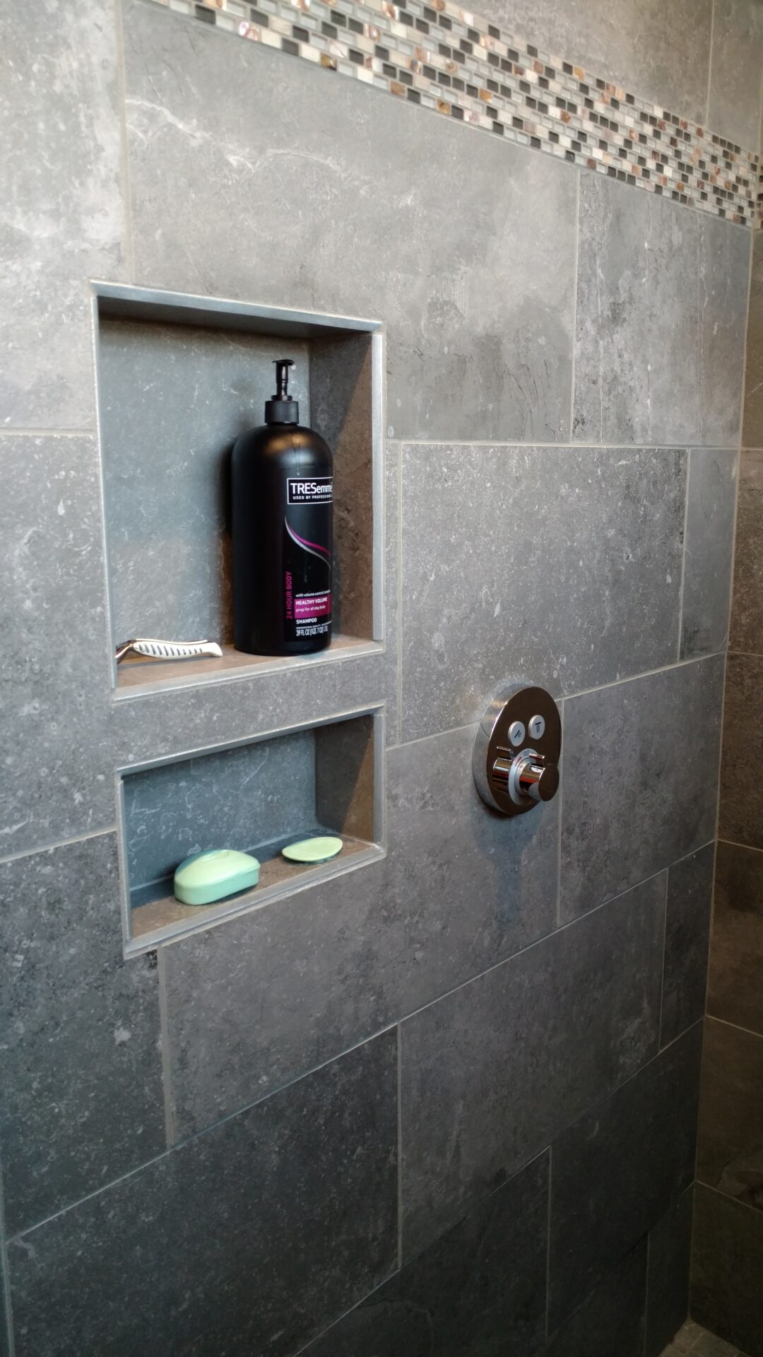 A tiled shower wall with built-in shelving, showcasing a black shampoo bottle, a comb, and two bars of soap. The wall is accented by a horizontal strip of small mosaic tiles, creating a visually appealing feature.