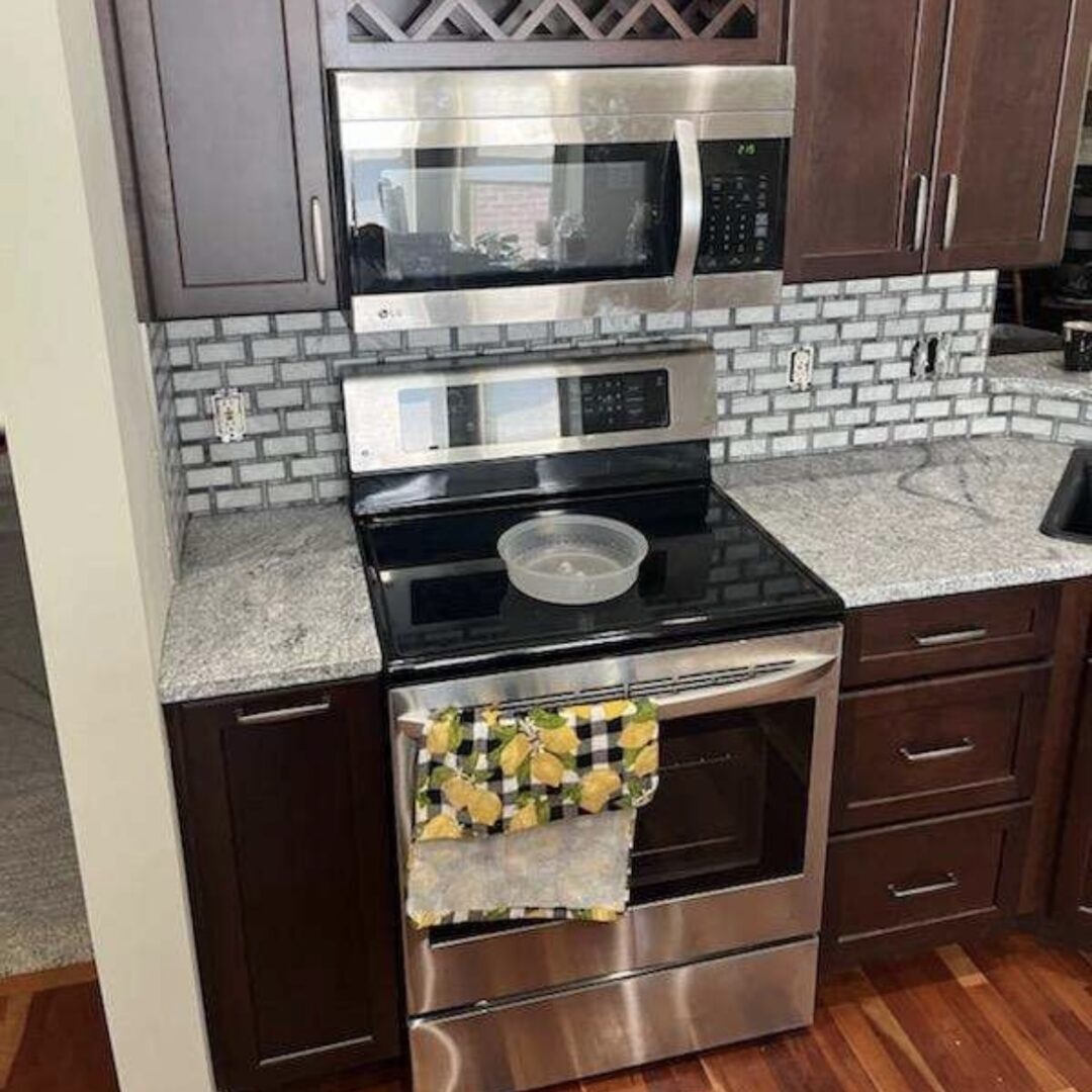 A kitchen with a modern stainless steel stove and microwave, bordered by dark wood cabinets and a granite countertop. A colorful lemon-print towel hangs from the oven, adding a cheerful accent to the space.